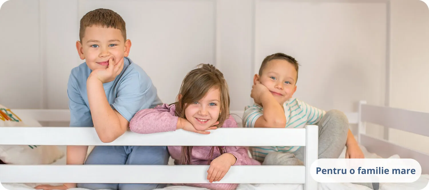 Trei copii zâmbitori sprijiniți de o barieră albă de pat.